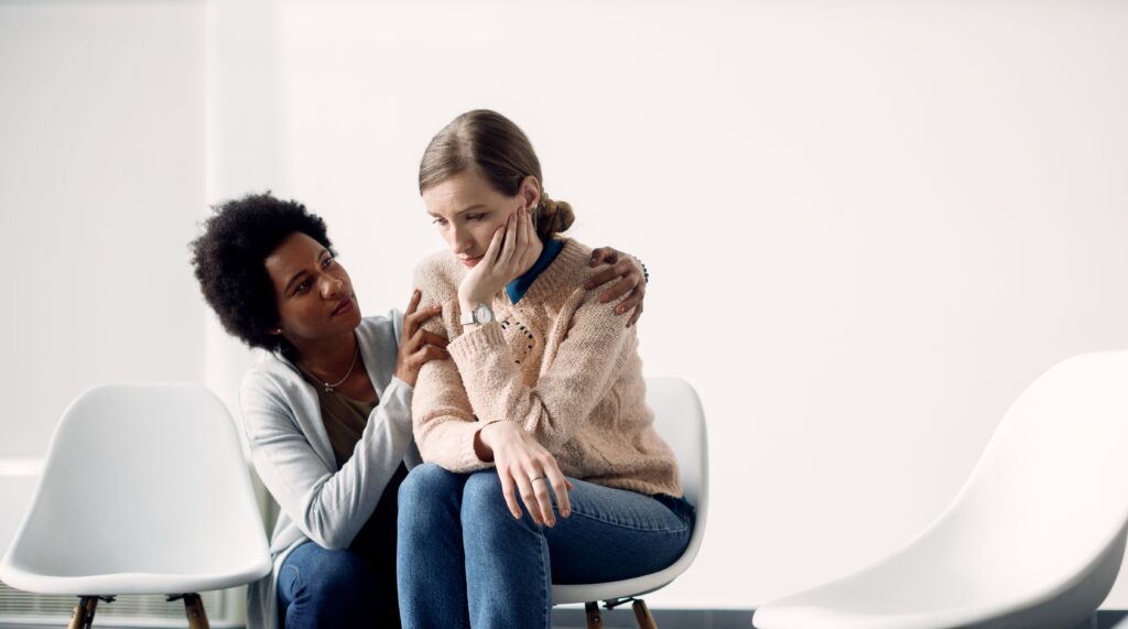 Duas mulheres sentadas em cadeiras, uma delas visivelmente triste e abatida, sendo consolada pela outra. A imagem representa apoio emocional e empatia, essenciais para superar traumas e ajudar quem está sofrendo.
