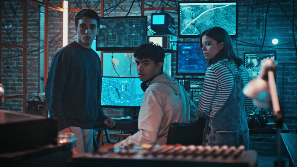 Três adolescentes olham assustados para trás como se estivessem sido surpreendidos por algo, eles estão em uma sala de controle tecnológica