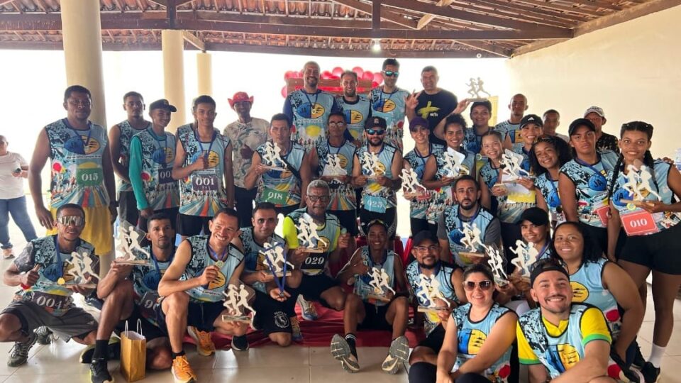 Grupo de pessoas reunidas em um espaço coberto, vestindo camisetas personalizadas com um design colorido e números de identificação. Muitos seguram troféus em formato de corredor, indicando uma premiação esportiva, possivelmente uma corrida. Todos sorriem e demonstram alegria pela conquista. O ambiente tem estrutura rústica, com pilares e telhado de madeira, e ao fundo há balões vermelhos decorativos.