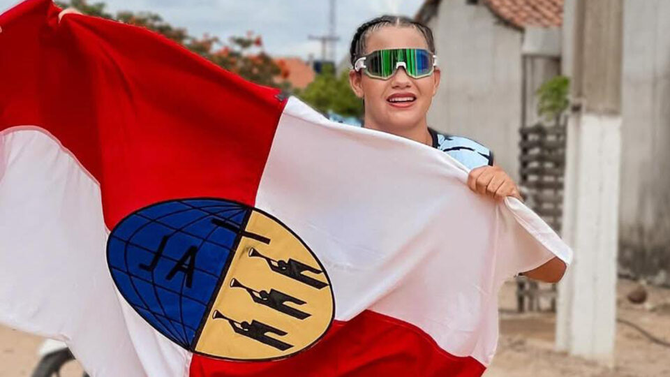 Mulher segurando uma bandeira vermelha e branca com um emblema ao centro, que contém um globo azul e figuras tocando trombetas. Ela veste óculos esportivos espelhados e uma camiseta azul, sorrindo enquanto olha para a câmera. O fundo mostra um ambiente externo com casas simples e uma bicicleta parcialmente visível.