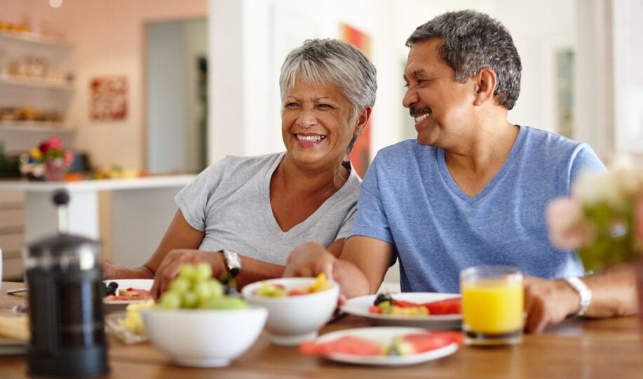 Casal de idosos felizes sentados à mesa tomando um café da manhã cheio de frutas para cuidar da saúde.