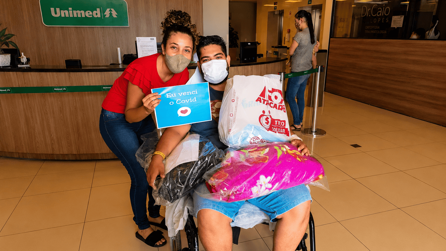 Andrew em uma cadeira de rodas saindo do hospital e Myllena próxima a ele segurando uma placa com o a frase "eu venci o COVID", demonstrando o cuidado de Deus com eles. Ambos de máscara.