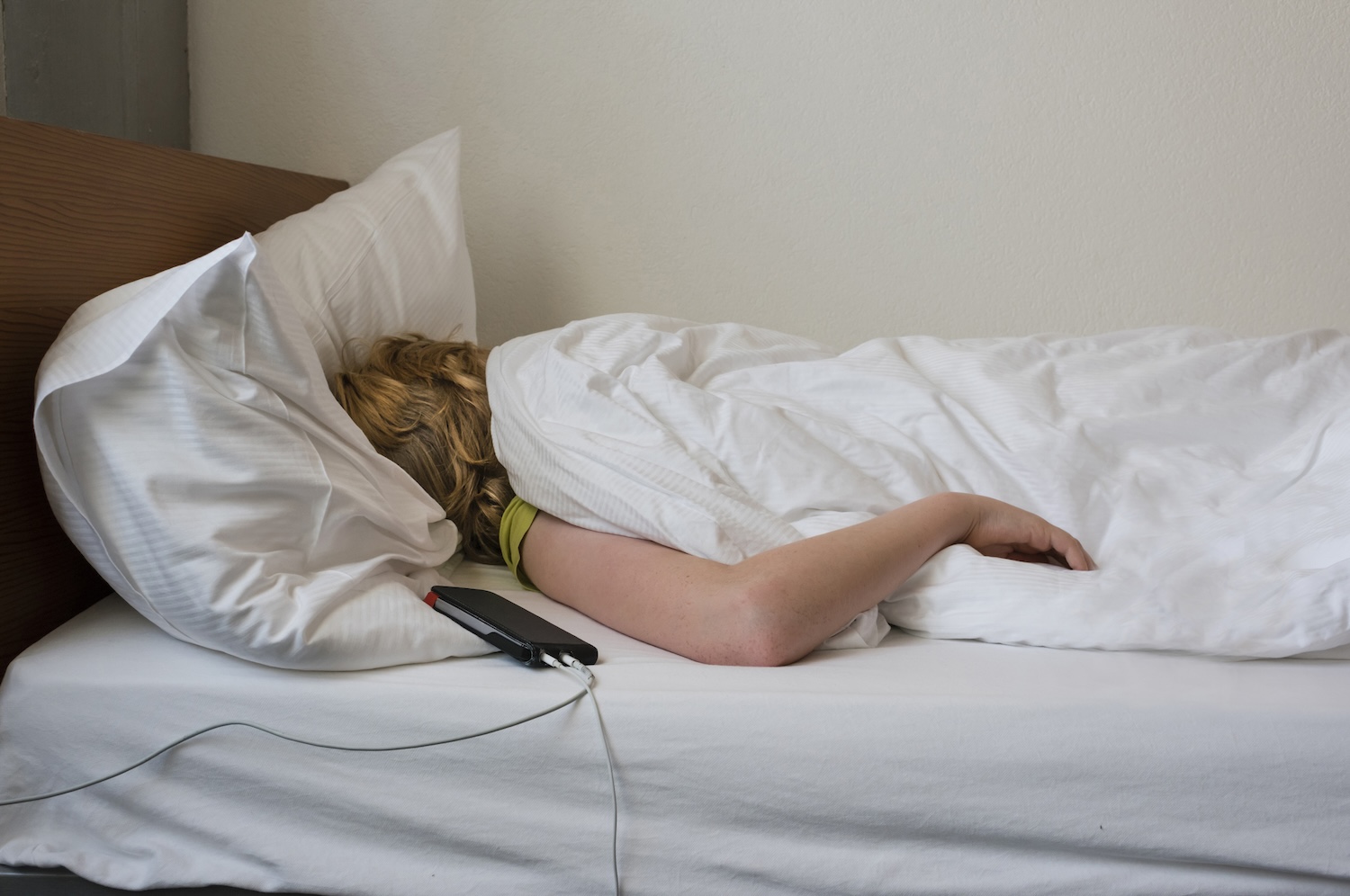 Adolescente está enrolado em cobertores durante um sono profundo em sua cama, ao lado de seu smartphone com fio e fones de ouvido.