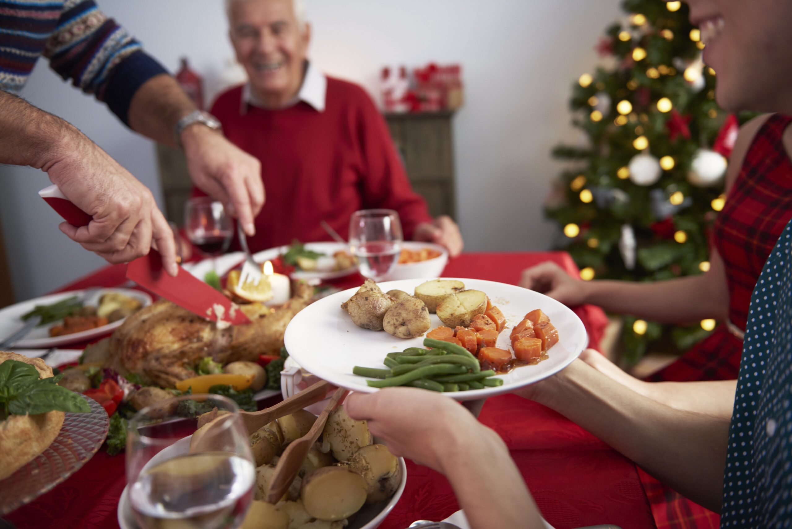 Família reunida em torno de uma mesa de jantar festiva, desfrutando de um banquete natalino. A imagem capta mãos passando um prato de comida, com destaque para um assado dourado, acompanhamentos de batatas, cenouras e vagem. Ao fundo, um idoso sorri, evidenciando a alegria do encontro. A decoração inclui uma toalha de mesa vermelha, velas acesas e um pinheiro de Natal iluminado ao fundo, transmitindo um ambiente acolhedor e festivo.