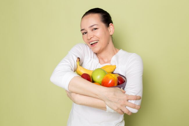 Mulher sorridente segurando uma tigela de frutas frescas, incluindo bananas, maçãs e uma pera, simbolizando uma alimentação saudável.