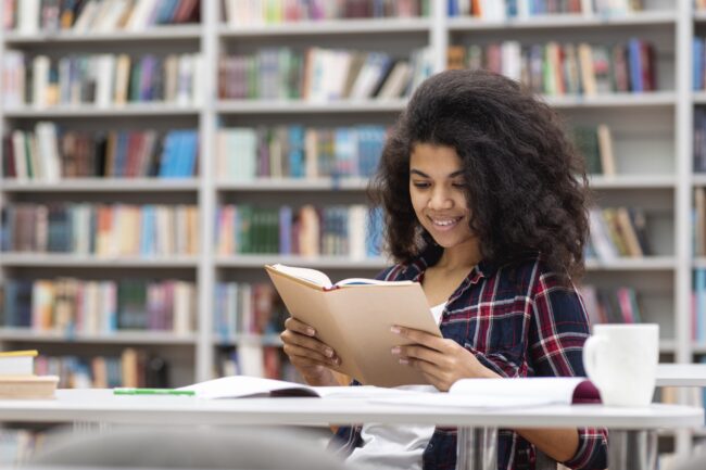 Mulher jovem sorrindo enquanto lê um livro em uma biblioteca, com prateleiras cheias de livros ao fundo.