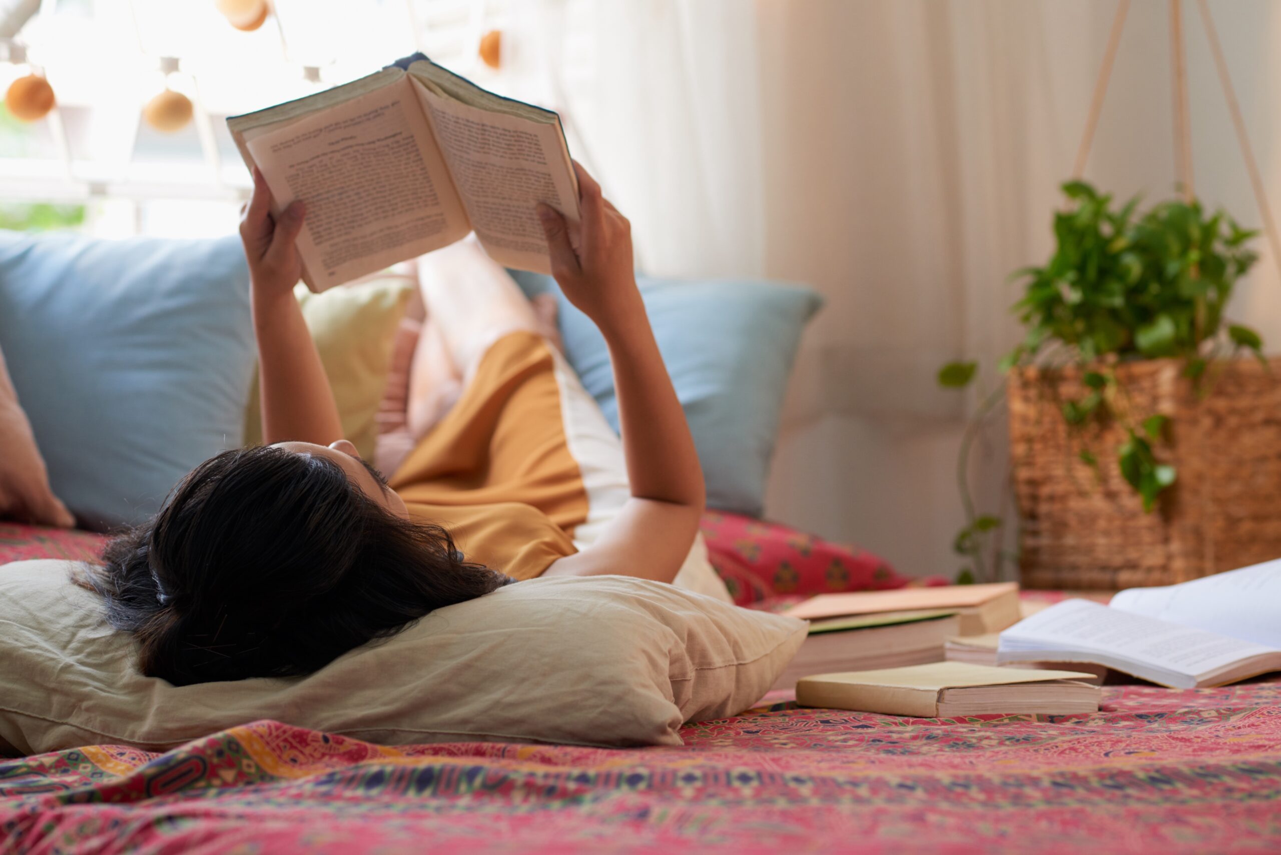 Pessoa deitada lendo um livro em um ambiente caseiro relaxante, cercada por plantas e luz natural.