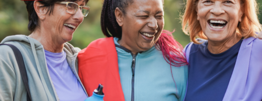 Três mulheres idosas felizes sorrindo e abraçadas ao ar livre, vestindo roupas esportivas coloridas, uma delas segurando uma garrafa de água. A imagem representa hábitos para uma vida saudável, associada ao momento vida e saúde.