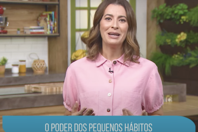 Mulher sorridente, vestindo uma camisa rosa, apresenta um programa em uma cozinha decorada, com o título "O Poder dos Pequenos Hábitos" exibido na parte inferior da imagem.
